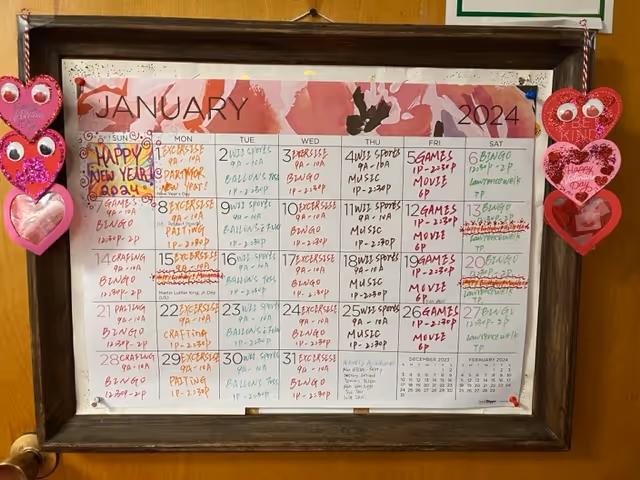 A framed January 2024 calendar hanging on a wooden wall, decorated with two red heart-shaped ornaments on either side. The calendar displays daily activities written in various colors, including exercise, games, bingo, music, and movies.