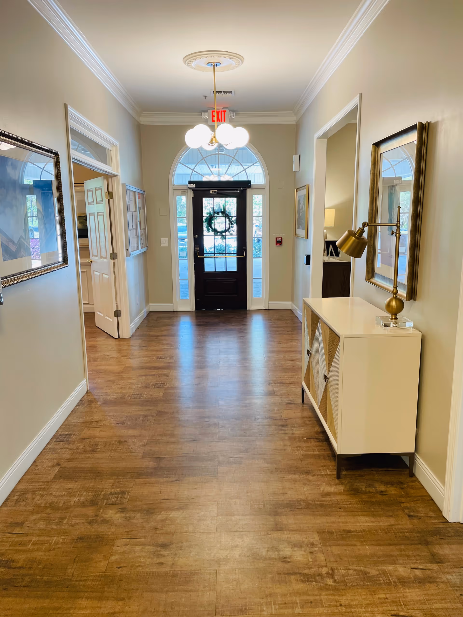 A well-lit hallway with wooden flooring leading to a glass door decorated with a wreath. The hallway has beige walls with white trim, a ceiling light fixture with multiple bulbs, a white cabinet with a gold lamp on the right side, and framed artwork on the walls. There are open doorways on both sides of the hallway.