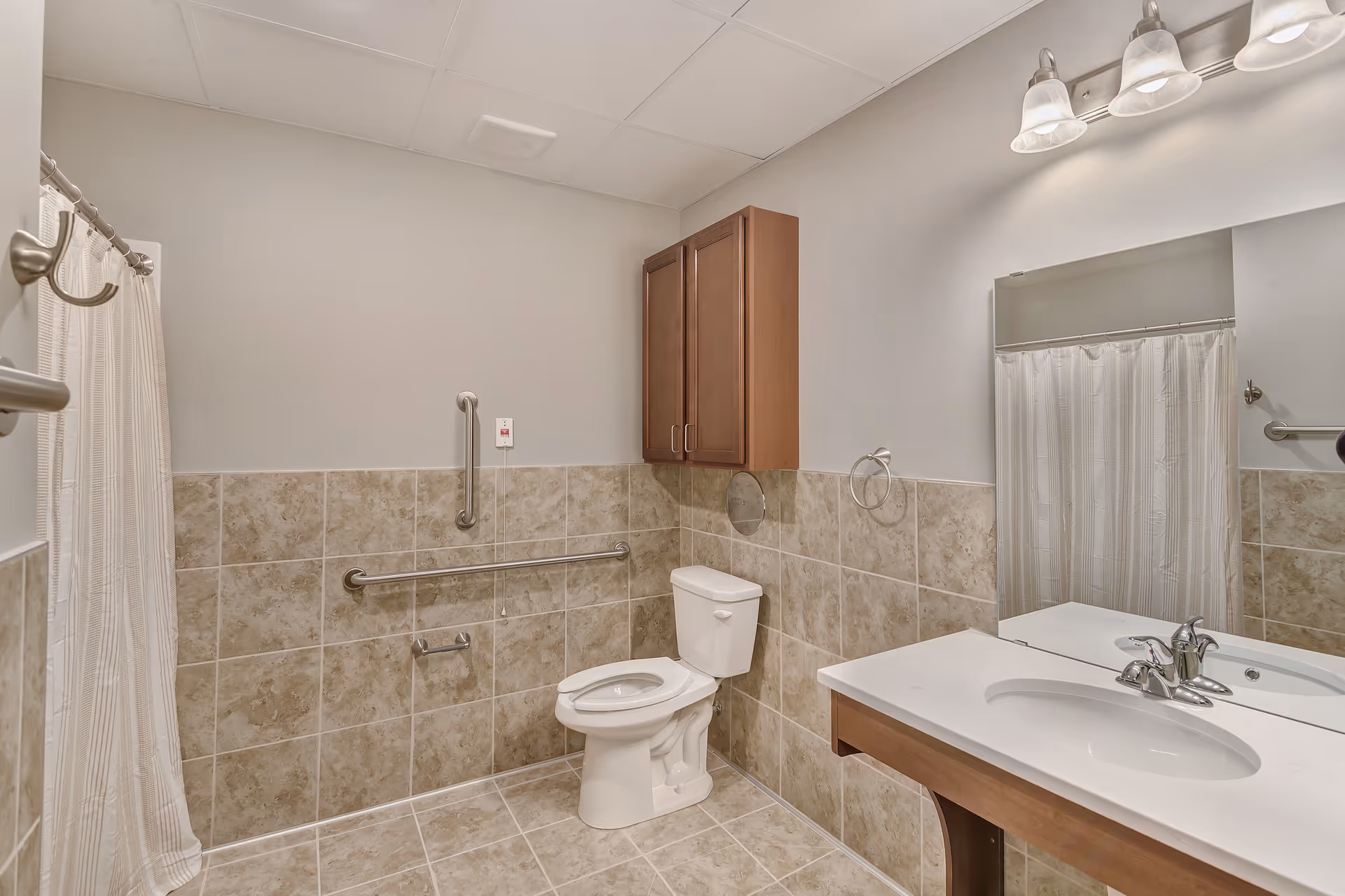 A clean and accessible bathroom with beige tiled walls and floor, a white toilet with grab bars on the wall, a wooden cabinet mounted above the toilet, a white sink with a wooden vanity, a large mirror, and a light fixture with three frosted glass shades. A shower curtain is partially visible on the left side.