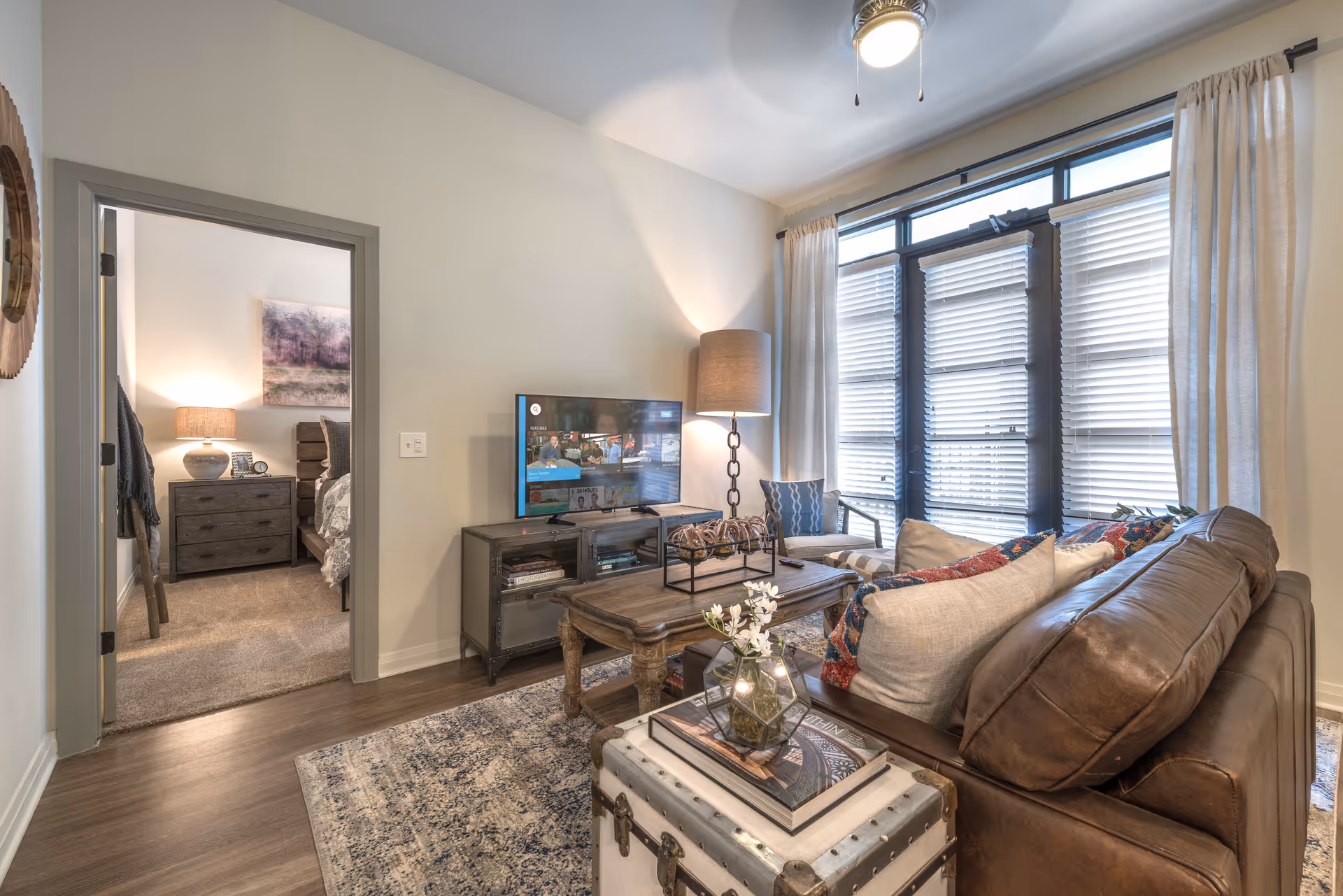 Cozy living room featuring a leather sofa, coffee table, TV stand, floor lamp, and large window with blinds, with a bedroom visible through an open doorway.