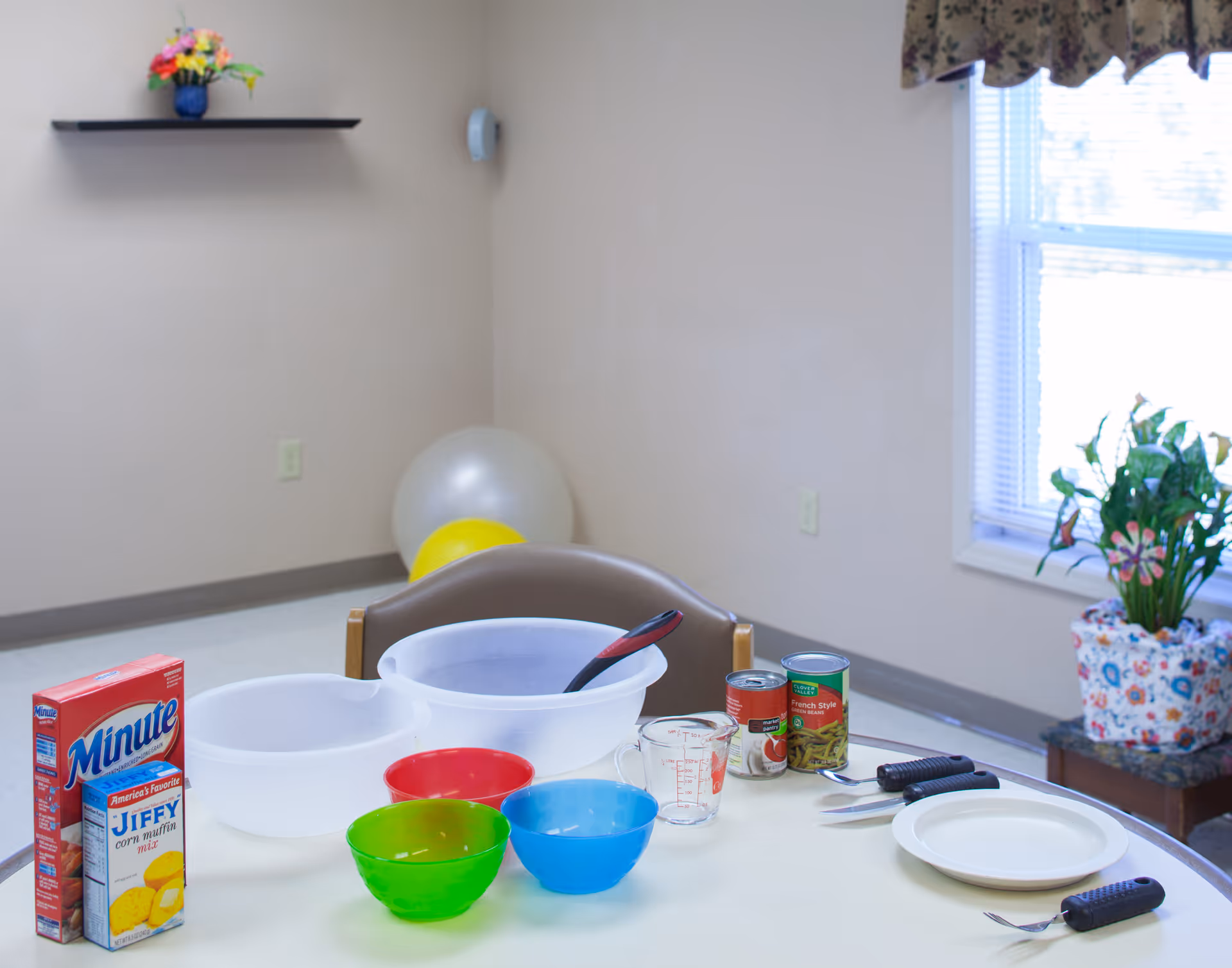 Table set with mixing bowls, colorful plastic bowls, canned ingredients, a measuring cup and utensils in a bright room with a window and potted plant.