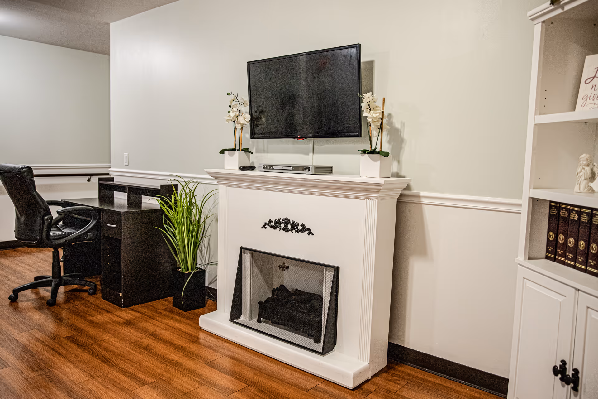 Interior room with a white electric fireplace topped by a flat-screen TV mounted on the wall. Two white orchid plants in square pots are placed on the fireplace mantel. To the left, there is a black office chair and a black desk with a green plant beside it. On the right, a white bookshelf holds books and decorative items. The floor is wooden, and the walls are painted light gray with white wainscoting.