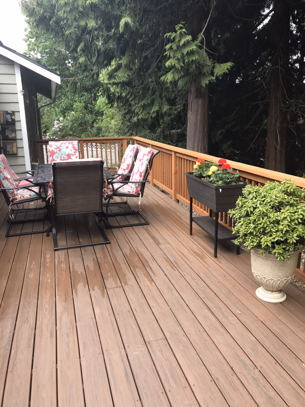 Outdoor wooden deck with a dining table and six chairs with floral cushions. There are two planters with flowers and greenery along the wooden railing. Tall trees and greenery surround the deck area.