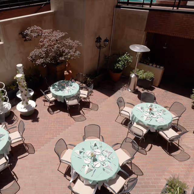 Outdoor patio area with round tables covered in light green tablecloths, each set with plates, glasses, and napkins. Several chairs surround each table. There are potted plants and decorative elements along the walls, and a tall outdoor heater is visible.