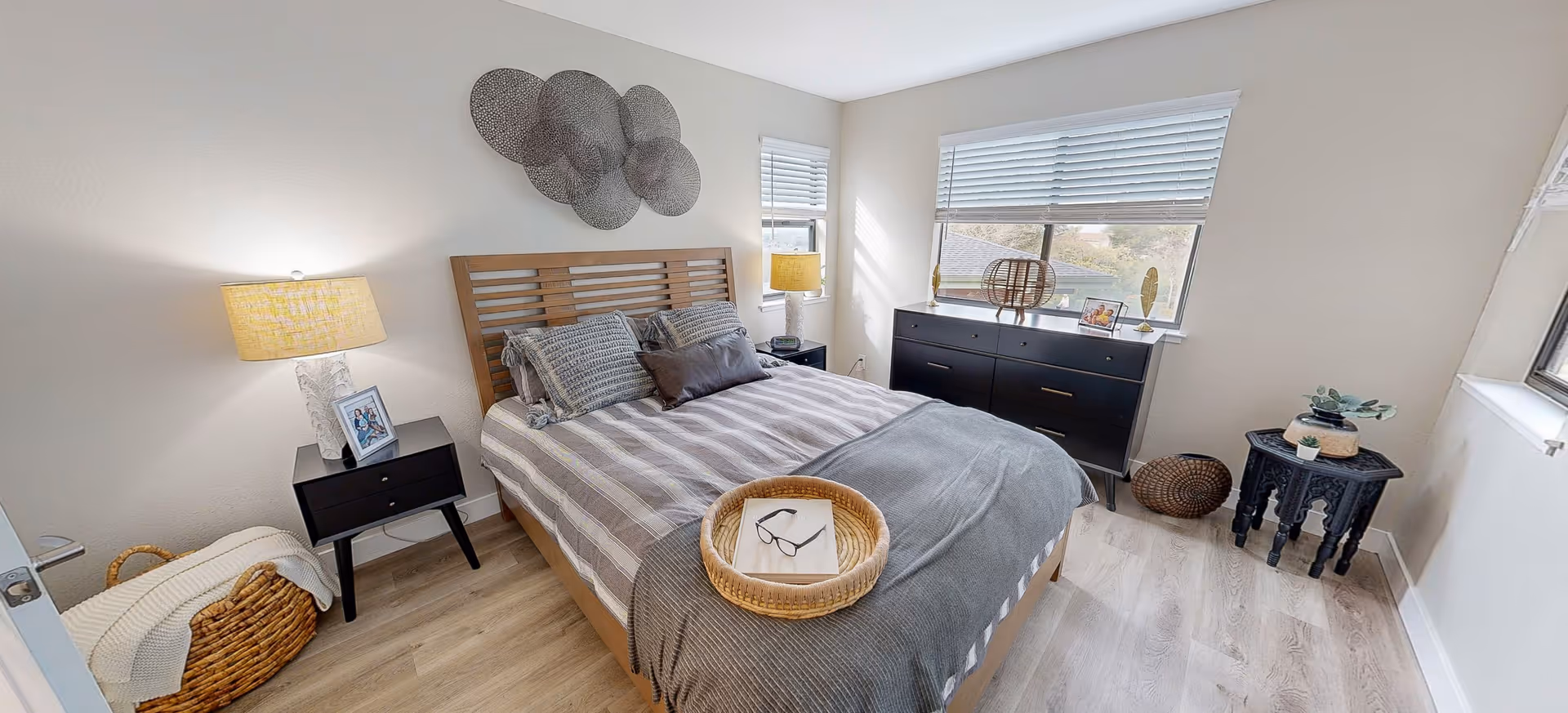 A bright and cozy bedroom with a wooden bed frame, striped bedding, and multiple pillows. There are two black nightstands with lamps on either side of the bed. A basket with a blanket is placed on the floor near the door. A black dresser is positioned under two windows with blinds, and decorative items are placed on top. The room has light-colored walls and wood flooring, with a small black side table and decorative pieces near the corner window.