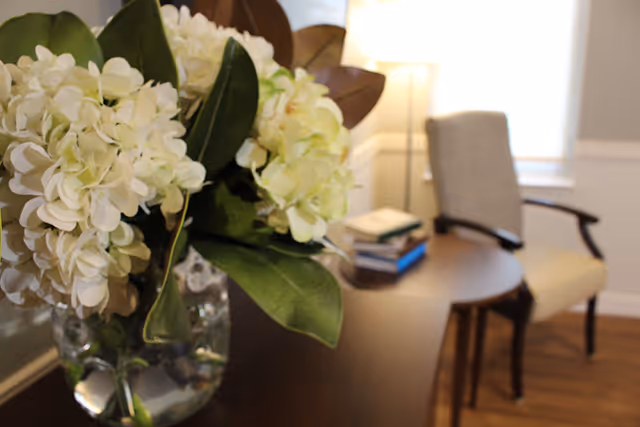 Close-up of a glass vase with white hydrangea flowers on a wooden table. In the background, there is a beige upholstered armchair, a small round wooden table with a few books on it, and a floor lamp near a window with white blinds.