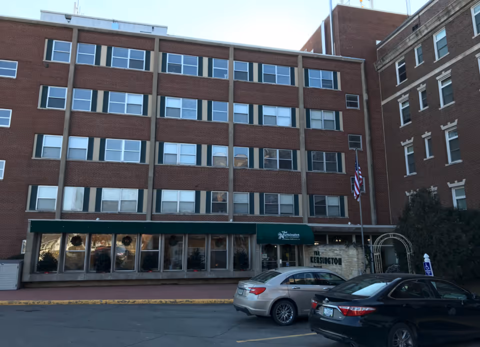 Brick four-story building with green awning, windows, and parked cars in front.