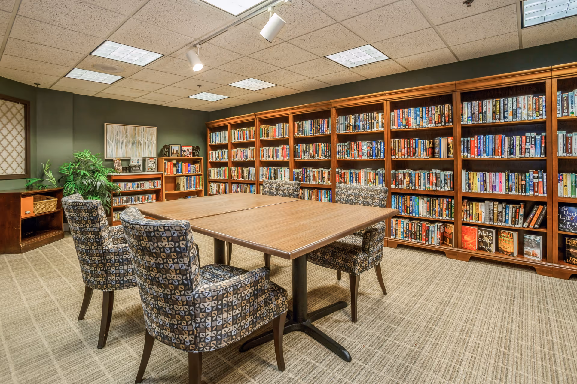 A cozy library room with wooden bookshelves filled with books along the walls. In the center, there is a wooden table with four patterned upholstered chairs around it. The room has a carpeted floor, green walls, and a ceiling with fluorescent lights and a spotlight. There is also a small plant and framed artwork on the wall.