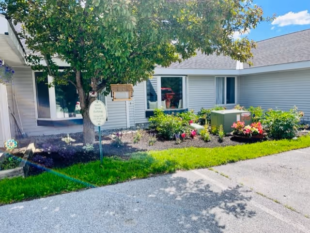 Front exterior of a single-story senior residence with windows, a tree, landscaped flower beds, a small sign, and adjacent parking.