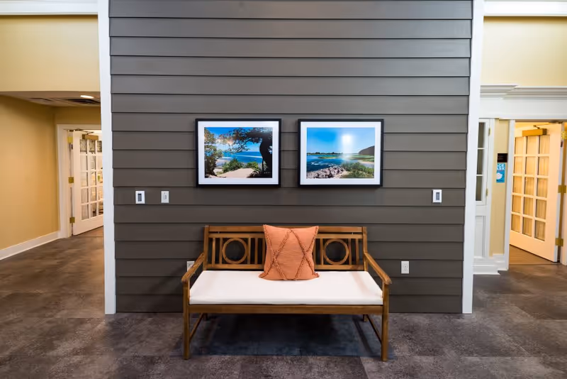 A wooden bench with a white cushion and an orange pillow is placed against a gray paneled wall. Above the bench, two framed landscape photographs are hung side by side. The room has a tiled floor and doorways on either side leading to other interior spaces with yellow walls and white trim.