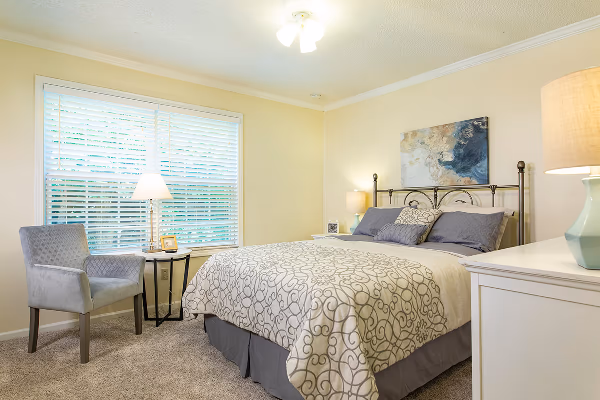 A cozy bedroom with a large bed featuring a decorative beige and gray patterned comforter and multiple pillows. There is a gray upholstered chair next to a small round side table with a lamp and a small framed picture. The room has beige walls, a large window with white blinds, a ceiling light fixture, and a piece of abstract artwork hanging above the bed. A white dresser with a lamp is partially visible on the right side.
