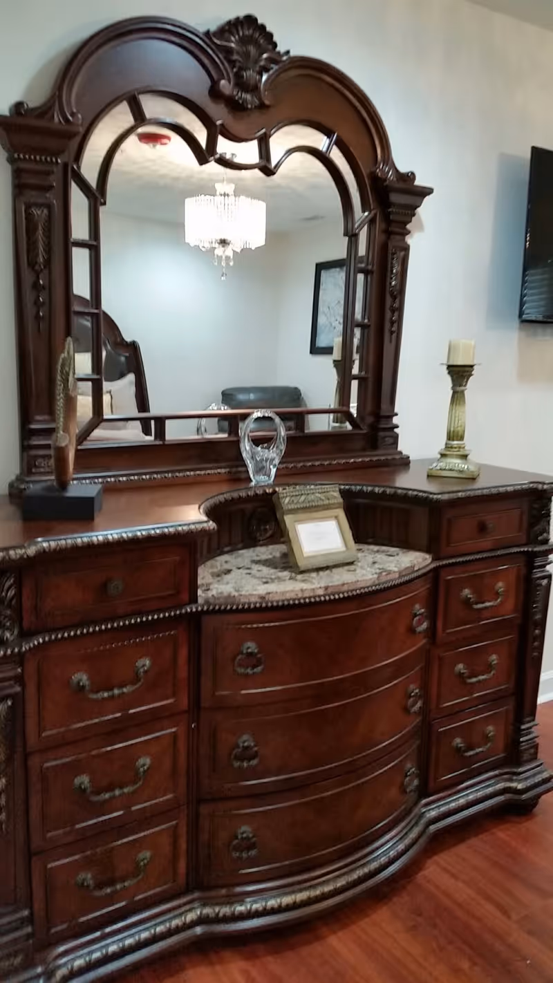 Ornate wooden dresser with multiple drawers and a large decorative mirror on top. The dresser has a marble inset and is adorned with a small framed picture, a glass decorative piece, a candle holder with a candle, and a feather ornament. The reflection in the mirror shows a chandelier, part of a chair, and a framed picture on the opposite wall.