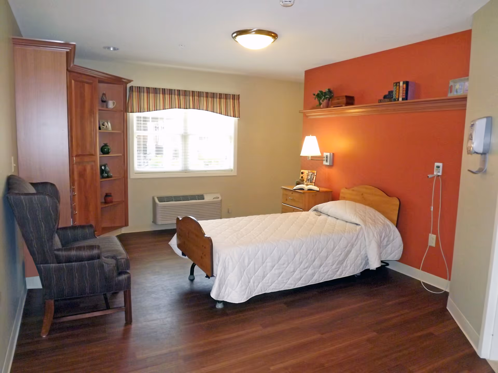 A furnished bedroom with a single bed, nightstand and armchair against an orange accent wall and a window letting in daylight.