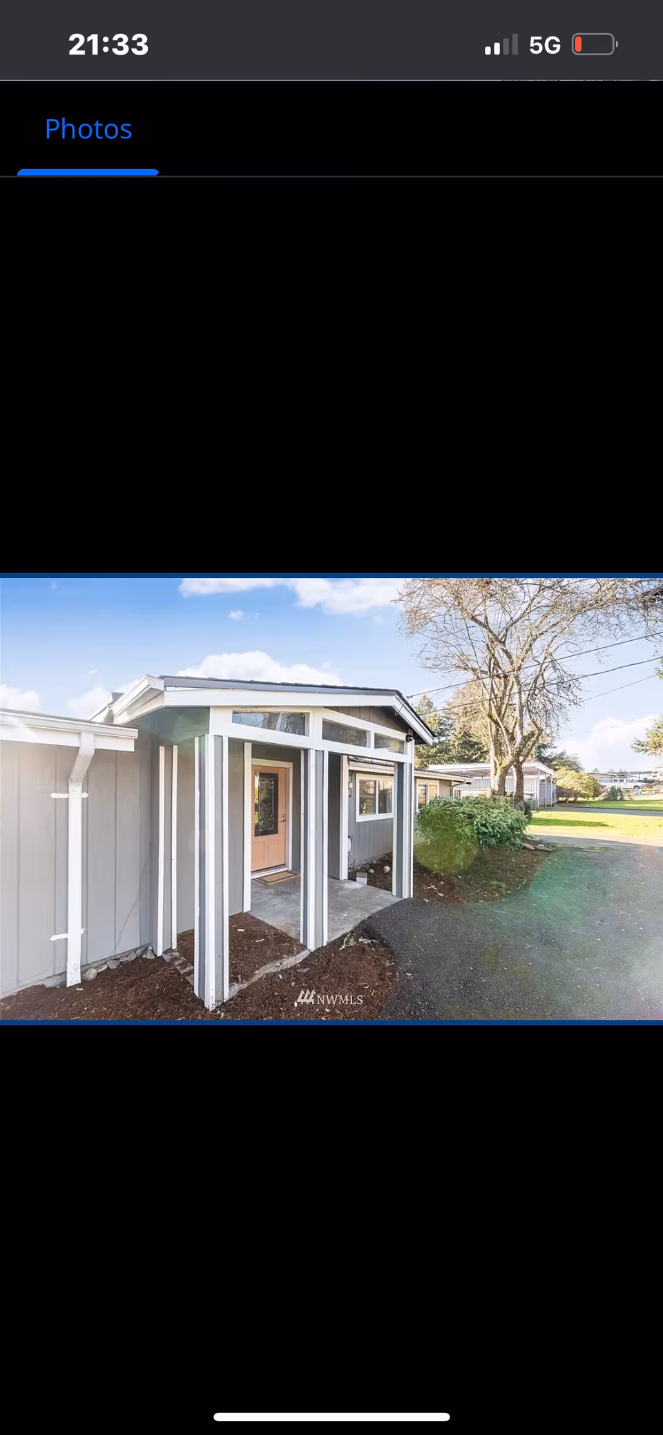 Exterior view of a single-story building entrance with a small covered porch supported by white pillars. The building is light gray with white trim, and there is a tree and green lawn visible to the right under a blue sky with some clouds.