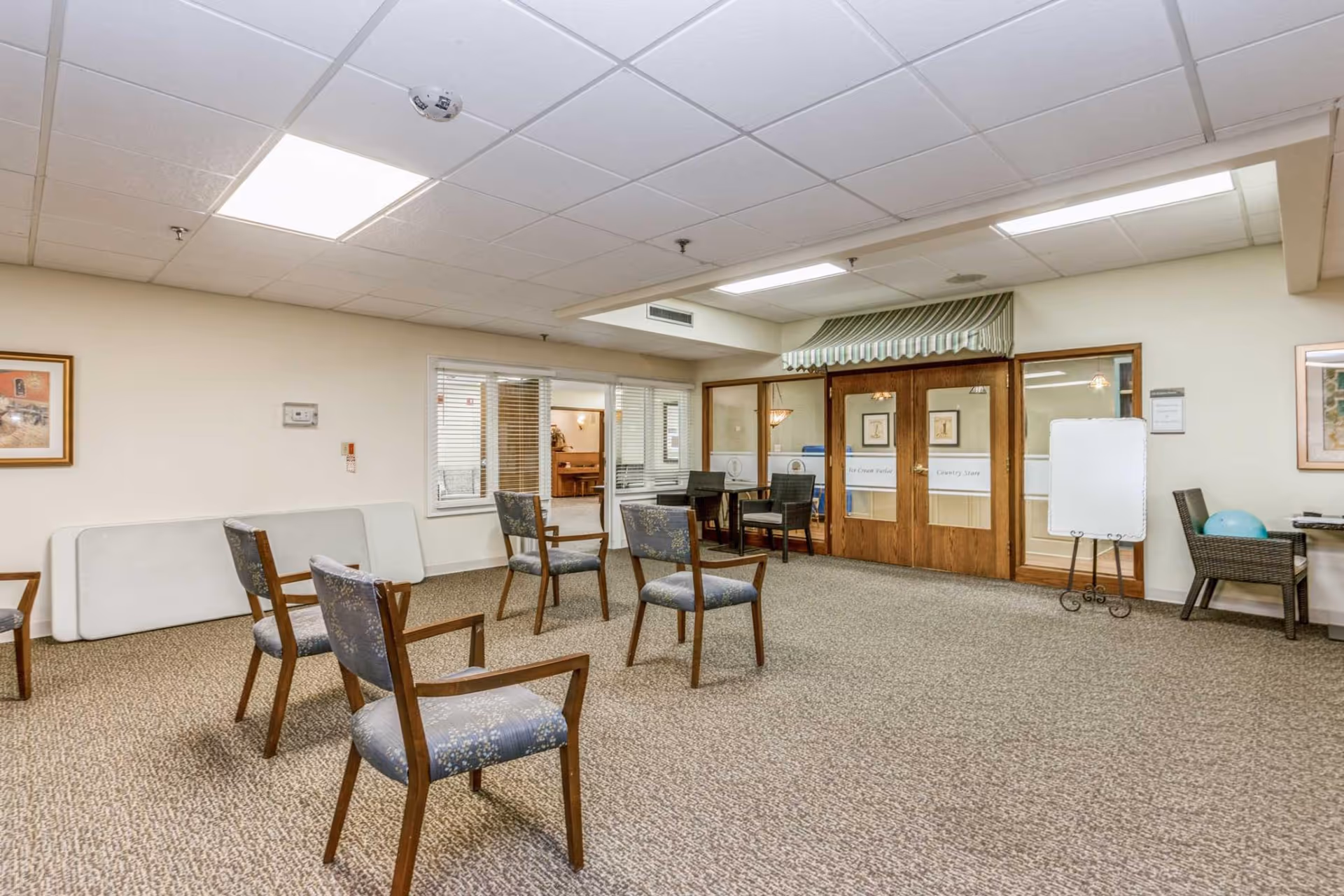 A spacious room with carpeted floor and several wooden chairs with patterned cushions arranged apart. The room has a drop ceiling with fluorescent lights. There are double wooden doors with glass panels labeled 'Ice Cream Parlor' and 'Country Store' with a green and white striped awning above. A small table with two chairs is near the doors, and a whiteboard on a stand is positioned to the right. The walls are light-colored with framed pictures hanging.