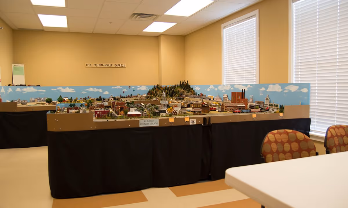 A room with a large model train layout titled 'The Fountainville Express' displayed on tables covered with black skirts. The model includes miniature buildings, trees, and a train track. The room has beige walls, two windows with white blinds, and patterned chairs near a white table.
