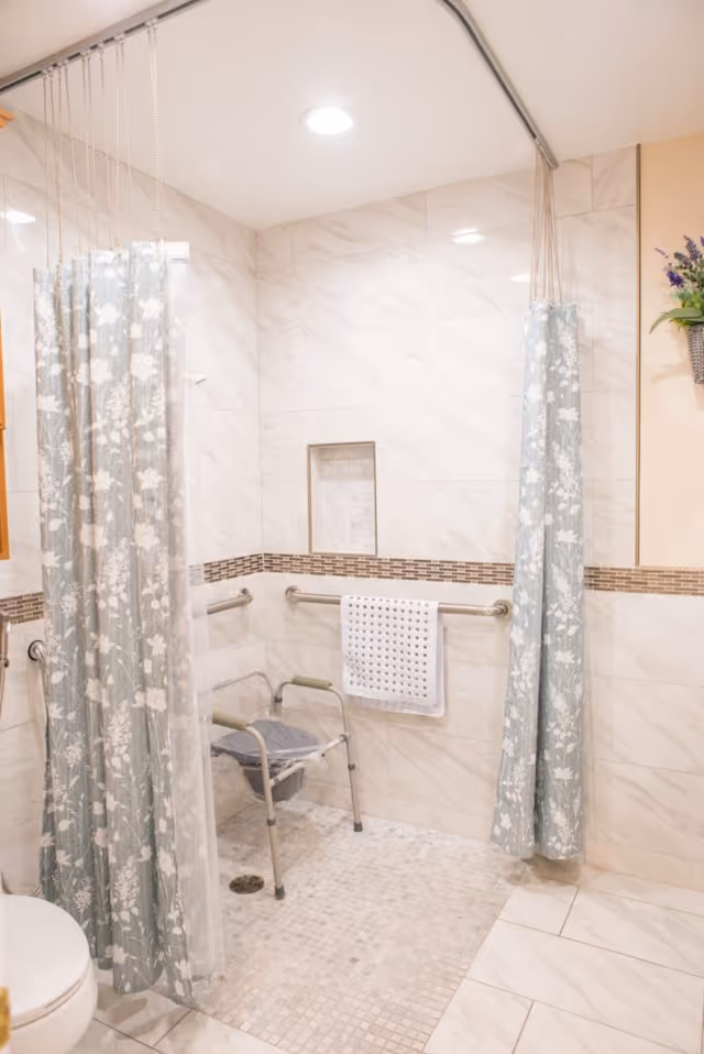 Accessible walk-in shower with floral curtains, grab bars, a shower chair, and tiled walls and floor.