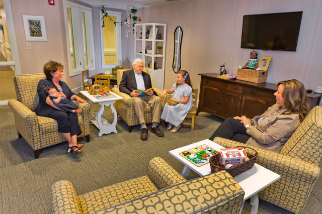 A group of residents and visitors sit and talk in a cozy assisted-living common room with armchairs, a TV, and children's toys.