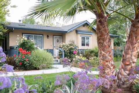 Single-story residential building with beige walls and blue trim, surrounded by lush greenery and colorful flowers including red, purple, and white blooms. Palm trees are visible in the foreground, and a concrete pathway leads to the front door.