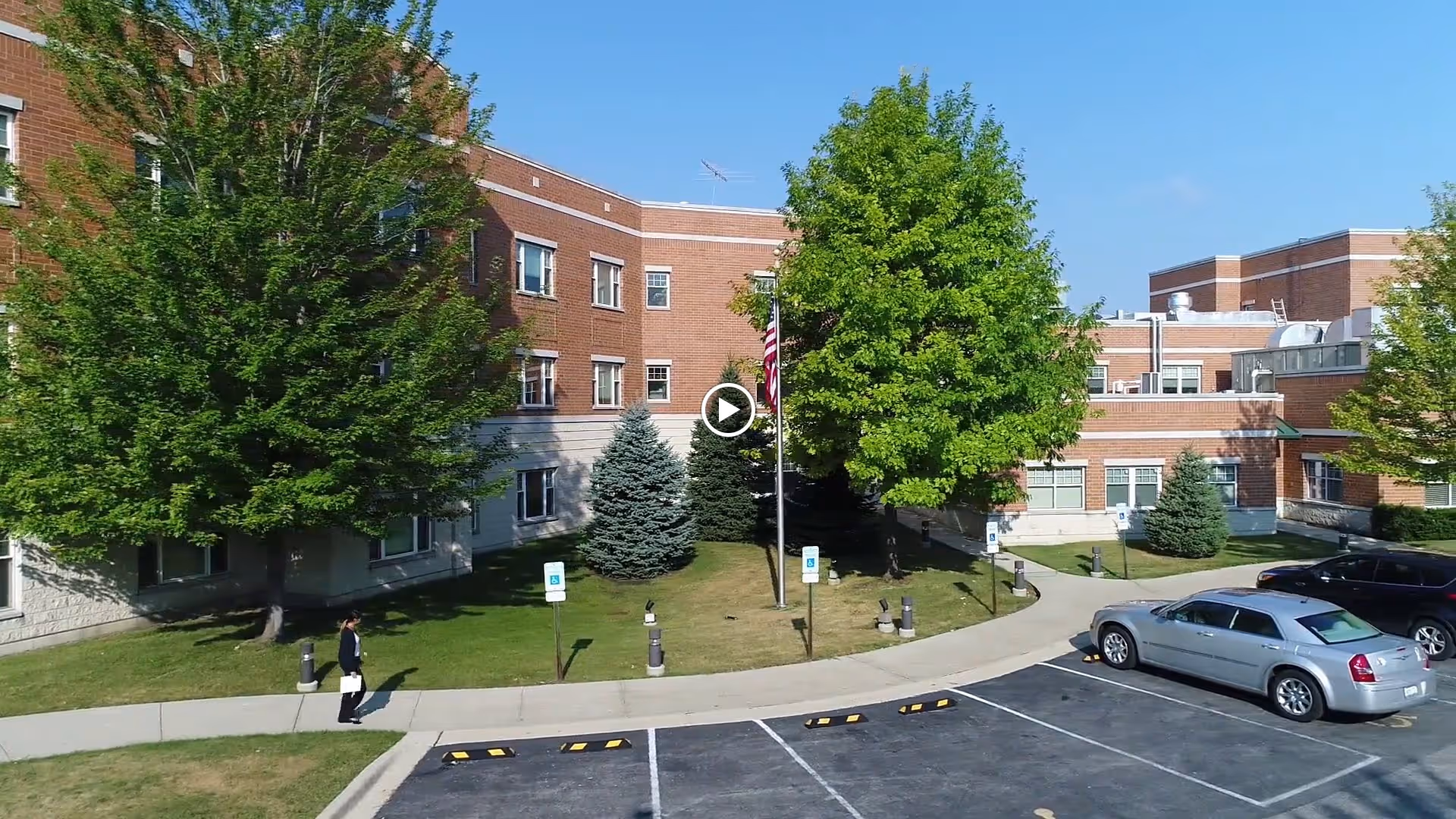 Front exterior of a brick senior living building with trees, a flagpole, walkway, and parked cars.