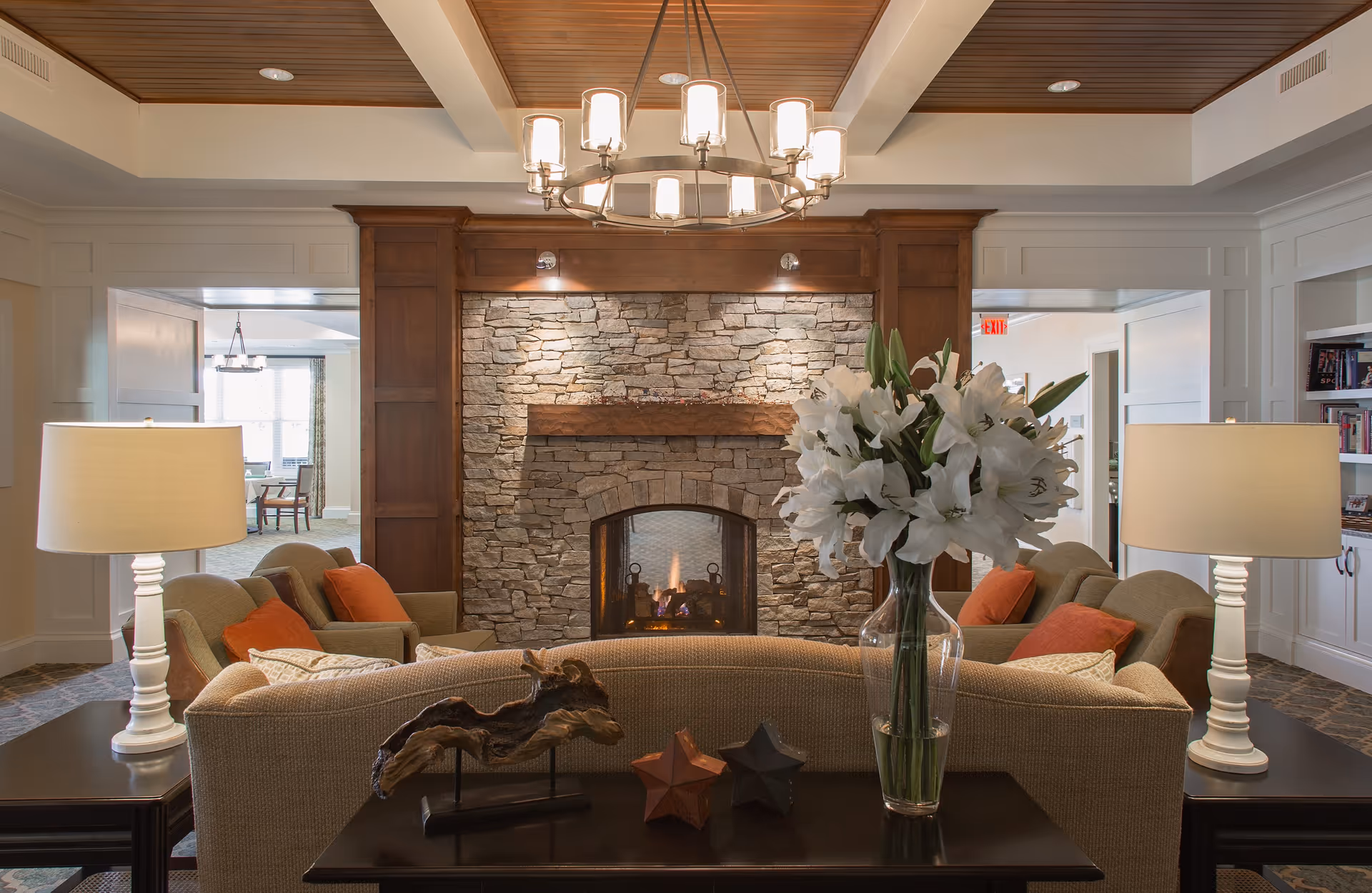 A cozy living room area with a stone fireplace in the center, flanked by wooden panels. In front of the fireplace is a beige sofa with orange and patterned cushions. On the table behind the sofa, there is a clear vase with white lilies, a piece of driftwood art, and two star-shaped decorations. Two white table lamps with beige shades are placed on side tables on either side of the sofa. The ceiling features wooden paneling and a modern chandelier with multiple lights. The room has an inviting and warm atmosphere.