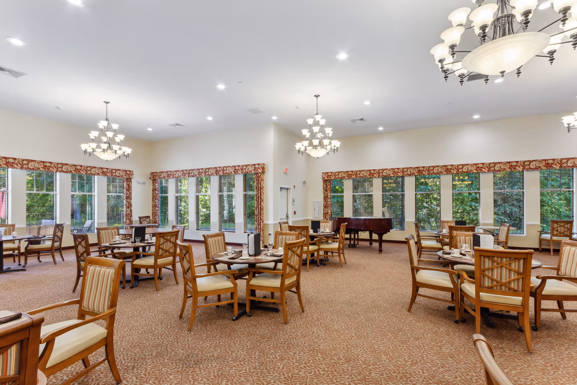 A spacious dining room with multiple round wooden tables and cushioned chairs arranged neatly. The room features large windows with floral valances, allowing natural light to brighten the space. Elegant chandeliers hang from the ceiling, and a grand piano is positioned near the windows in the background.