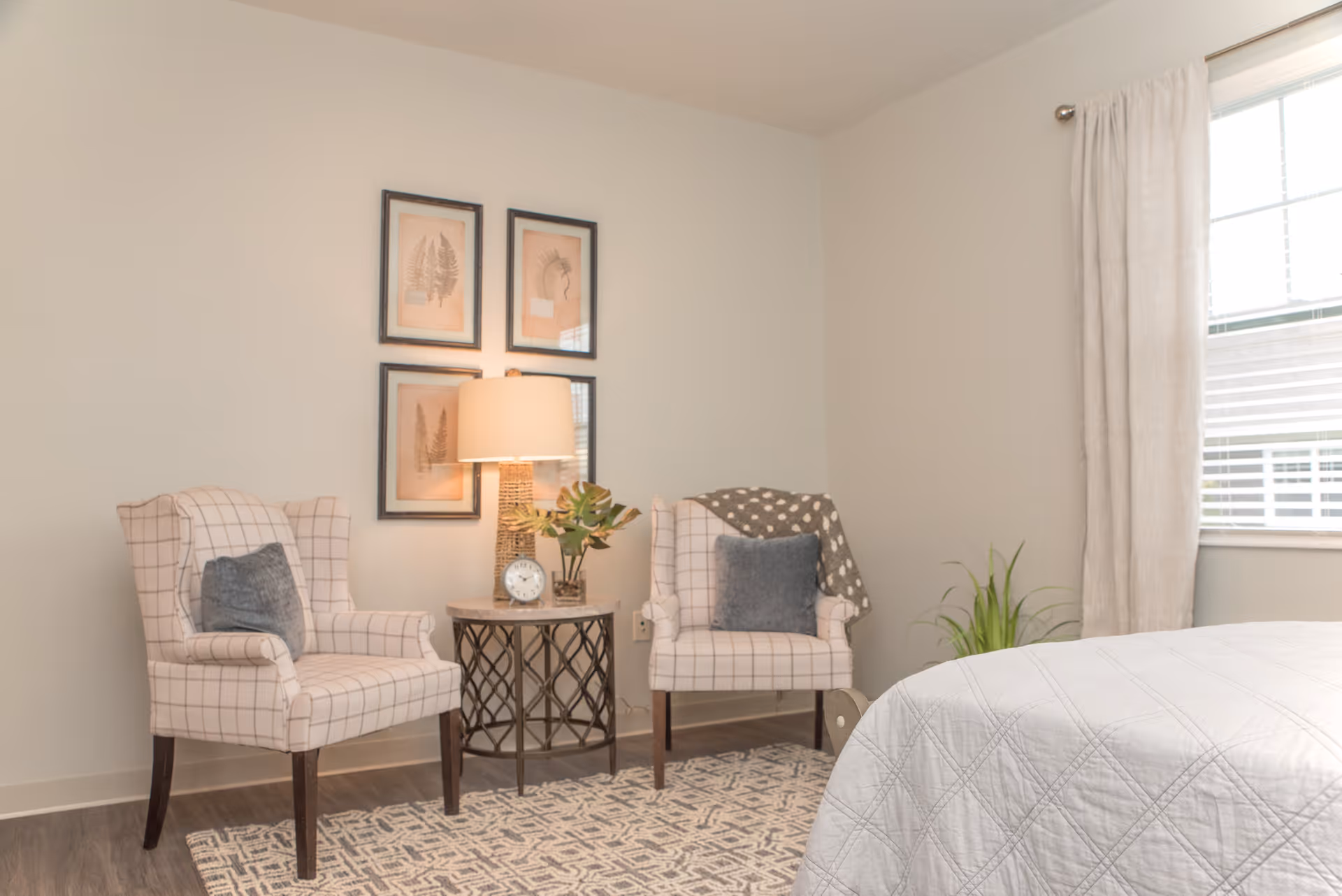 A cozy bedroom corner with two patterned armchairs, a small round table with a lamp, a clock, and a plant. Three framed botanical prints hang on the wall above the table. A bed with a white quilt is partially visible on the right side near a window with white curtains.