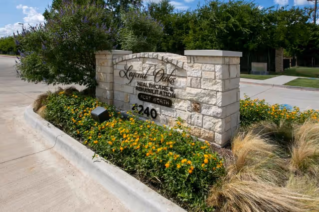 Stone sign for Legend Oaks Healthcare & Rehabilitation Fort Worth surrounded by green bushes and yellow flowers, located next to a concrete driveway under a blue sky with some clouds.