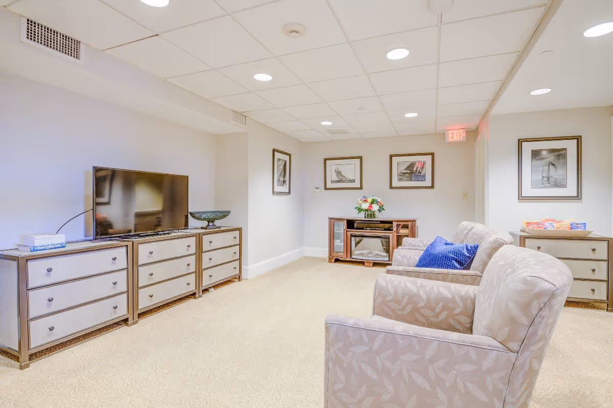 A cozy living room area in a senior living facility with beige patterned armchairs, a TV on a dresser, framed pictures on the walls, a small cabinet with a flower vase, and a side table with snacks. The room has a light-colored carpet and a drop ceiling with recessed lighting.