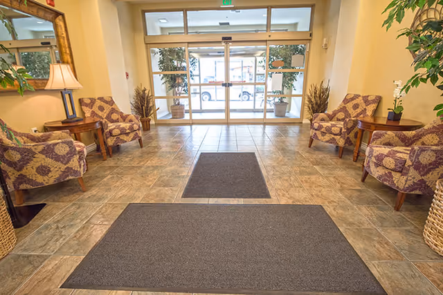 Entrance lobby area with tiled floor, two large gray mats, four patterned armchairs arranged in pairs on either side, wooden side tables with lamps and plants, large mirror on the left wall, and glass double doors leading outside with potted plants visible through the doors.