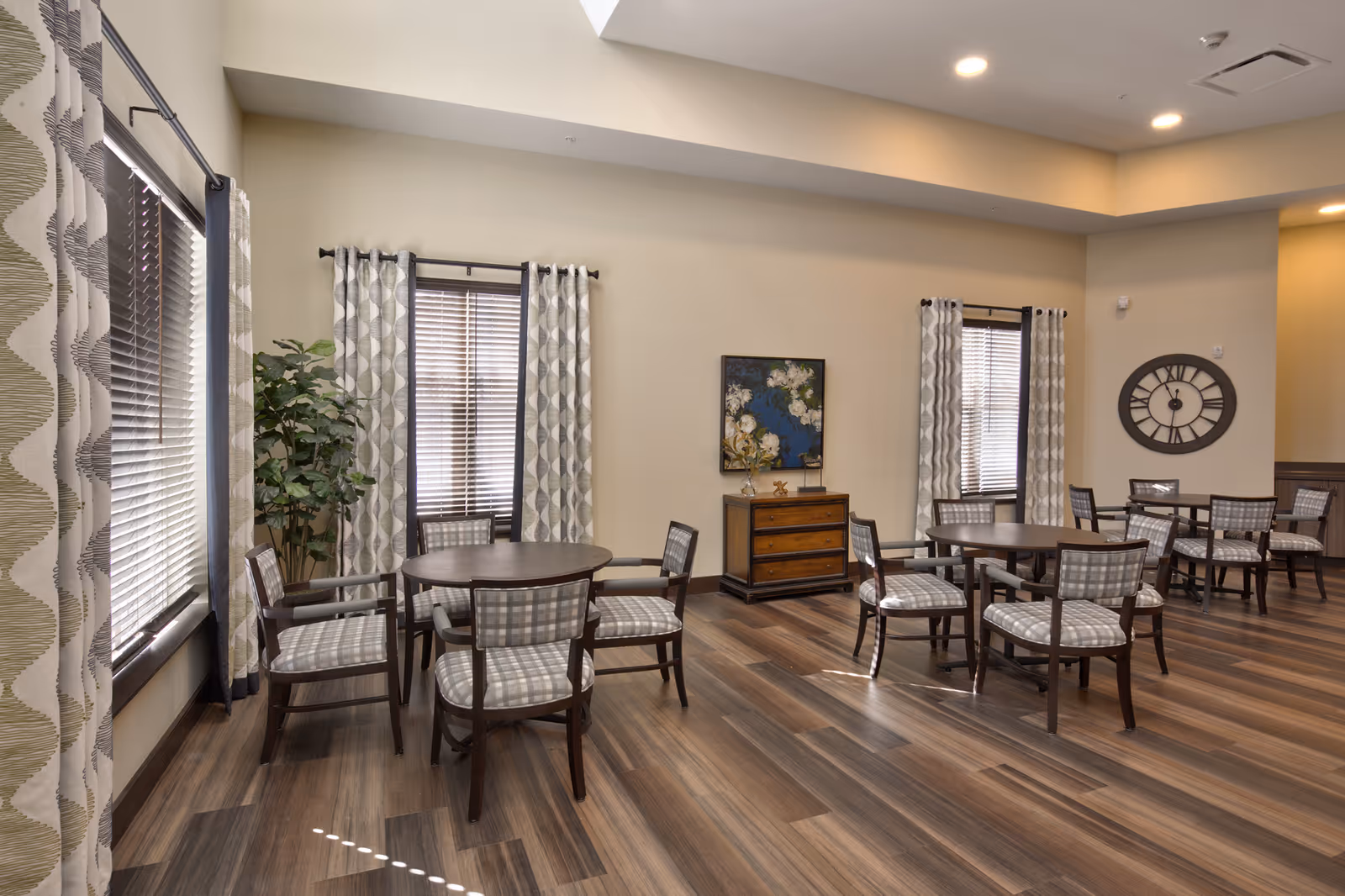 A spacious, well-lit common area with several round wooden tables surrounded by cushioned chairs with patterned upholstery. The room features large windows with patterned curtains, a wooden floor, a decorative plant, a chest of drawers with a floral painting above it, and a large wall clock.
