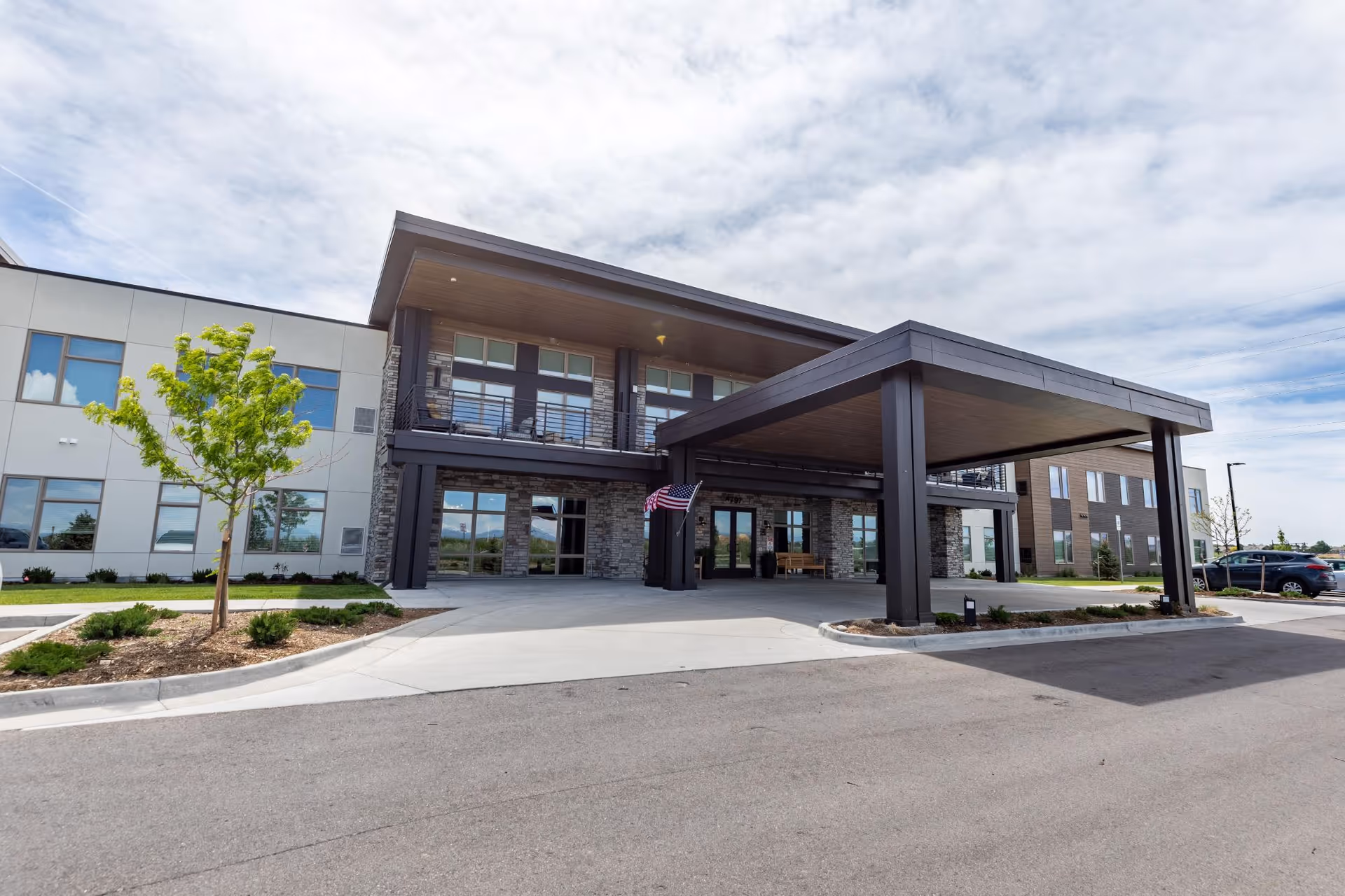 Exterior front view of a modern senior living facility building with a covered entrance, large windows, a small tree, and a few parked cars under a partly cloudy sky.