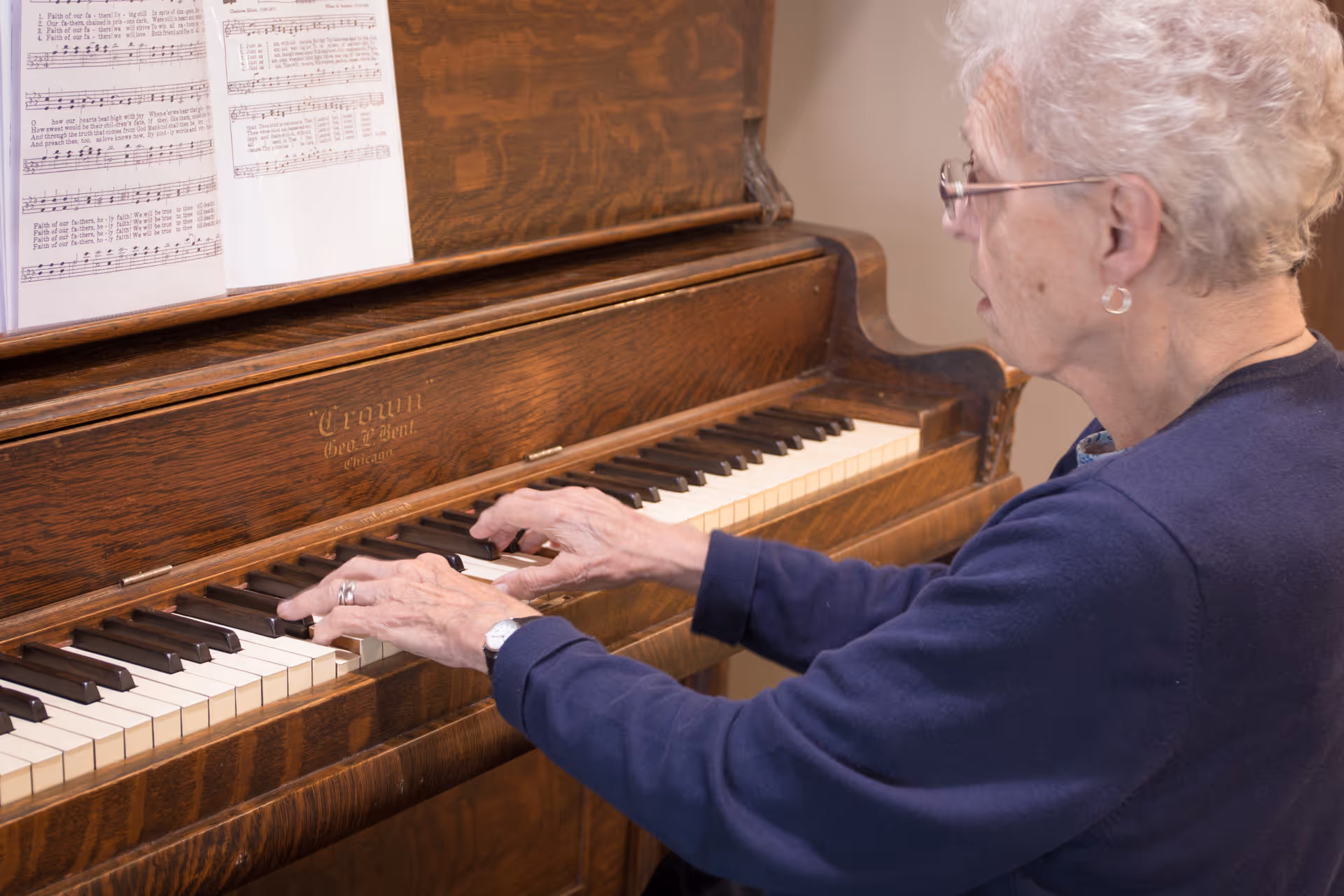 An elderly woman with short white hair and glasses is playing an upright wooden piano with sheet music placed on the music stand.