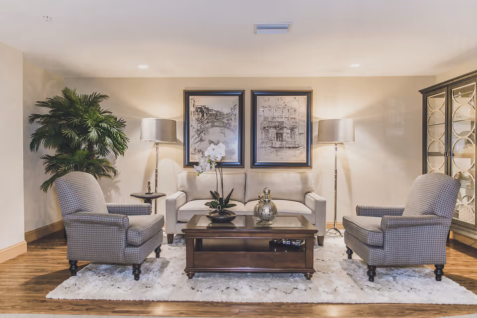 A cozy living room area with a beige sofa centered between two patterned armchairs. A dark wooden coffee table with decorative items sits on a white fluffy rug. Two floor lamps flank the sofa, and two framed black-and-white artworks hang on the wall behind it. A large green plant is on the left side, and a glass-front cabinet is on the right.