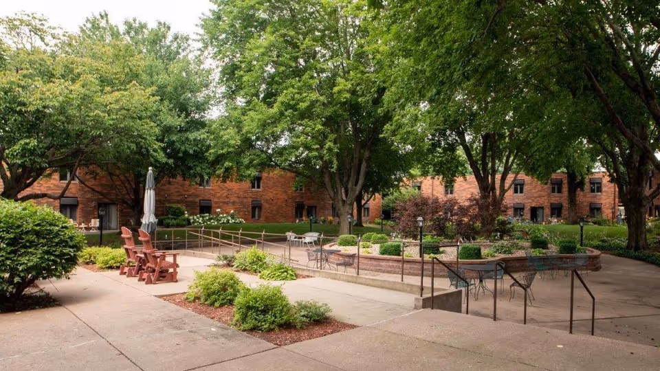 Outdoor courtyard area at Ridgecrest Village featuring concrete walkways, several trees providing shade, landscaped bushes, and a circular raised garden bed with plants. There are multiple seating areas with metal tables and chairs, as well as wooden Adirondack chairs and a closed umbrella.