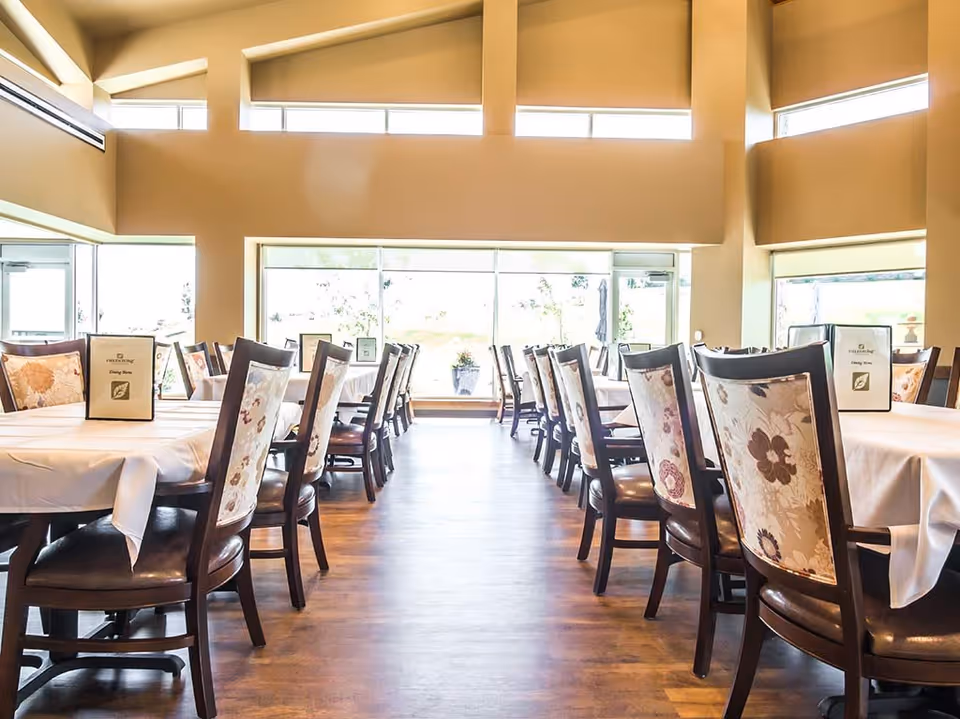 Bright dining room with multiple tables covered in white tablecloths and chairs with floral patterned backs. Large windows let in natural light, and the room has high ceilings with beige walls.