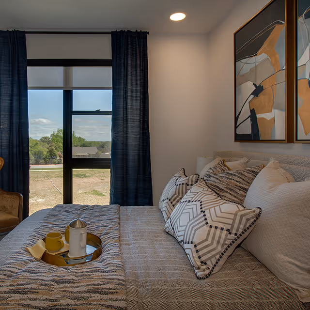 A cozy bedroom with a neatly made bed featuring multiple patterned pillows and a textured blanket. A tray with a yellow cup and a white container sits on the bed. The room has a large window with dark blue curtains, allowing natural light to brighten the space. Two abstract paintings hang on the wall above the bed.