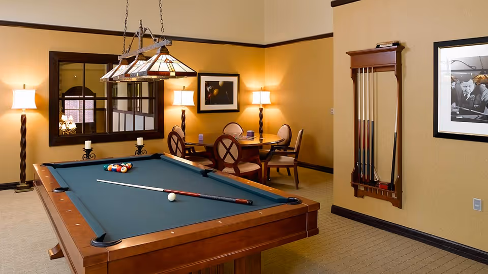 A cozy game room with a pool table in the center, a cue stick resting on the table, and a rack of pool cues mounted on the wall. There is a round table with four chairs in the background, two floor lamps, a large mirror, and framed artwork on the walls. The room has warm lighting and beige walls with dark wood trim.
