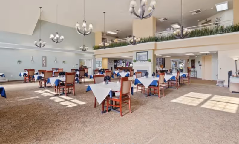 A spacious dining room in a senior living facility with multiple tables covered with white and blue tablecloths, wooden chairs, chandeliers hanging from the ceiling, and large windows allowing natural light to fill the room.