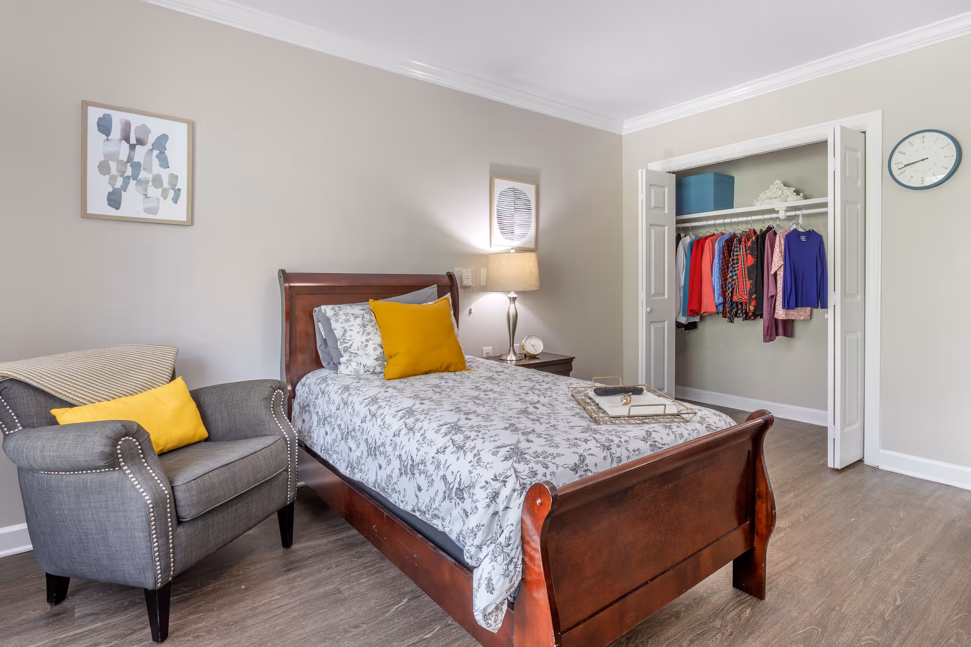 A cozy bedroom with a wooden bed featuring floral bedding and a yellow accent pillow. Next to the bed is a gray armchair with a yellow pillow and a beige throw blanket. A nightstand with a lamp and a clock is beside the bed. The closet doors are open, showing hanging clothes and storage boxes. The room has light-colored walls, a framed abstract artwork, and a wall clock.