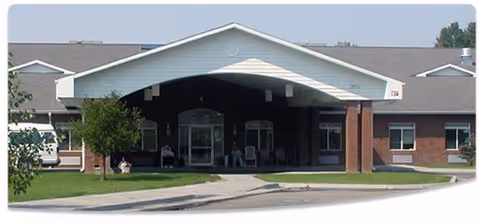 Front entrance of a single-story assisted living facility with a covered porte-cochere, brick facade, driveway and lawn.