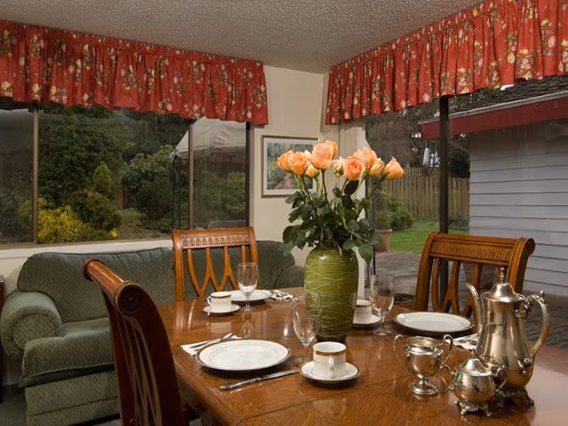 A dining room with a wooden table set for four, including plates, cups, glasses, and silverware. A green vase with peach-colored roses is in the center of the table. Behind the table is a green upholstered couch and large windows with red floral valances, showing a garden outside.