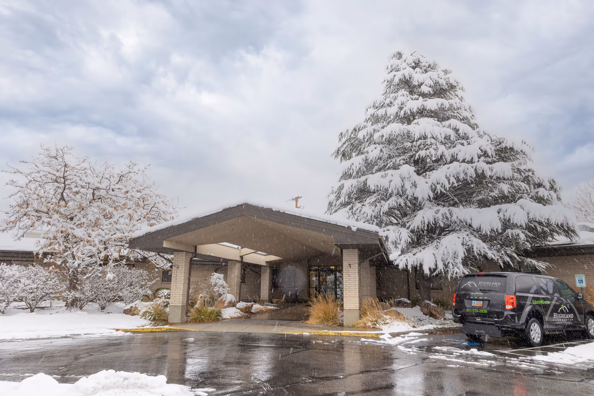 Snow-covered entrance of Highland Care Center with a large tree and bushes also covered in snow. A black vehicle with Highland Care Center branding is parked near the entrance under a cloudy sky.