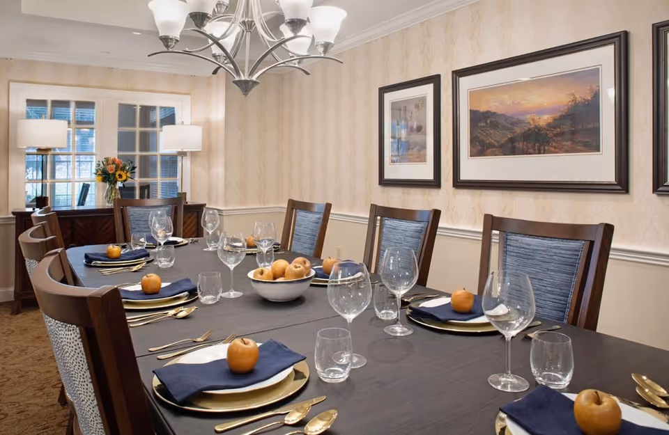 A formal dining room with a long dark wooden table set for eight people. Each place setting includes a gold charger plate, white plate, navy blue napkin, gold cutlery, a wine glass, and a water glass. Small apples are placed on each napkin. A bowl of apples is in the center of the table. The room has beige wallpaper, framed landscape paintings on the wall, a chandelier overhead, and two table lamps on a sideboard near a window with white trim.