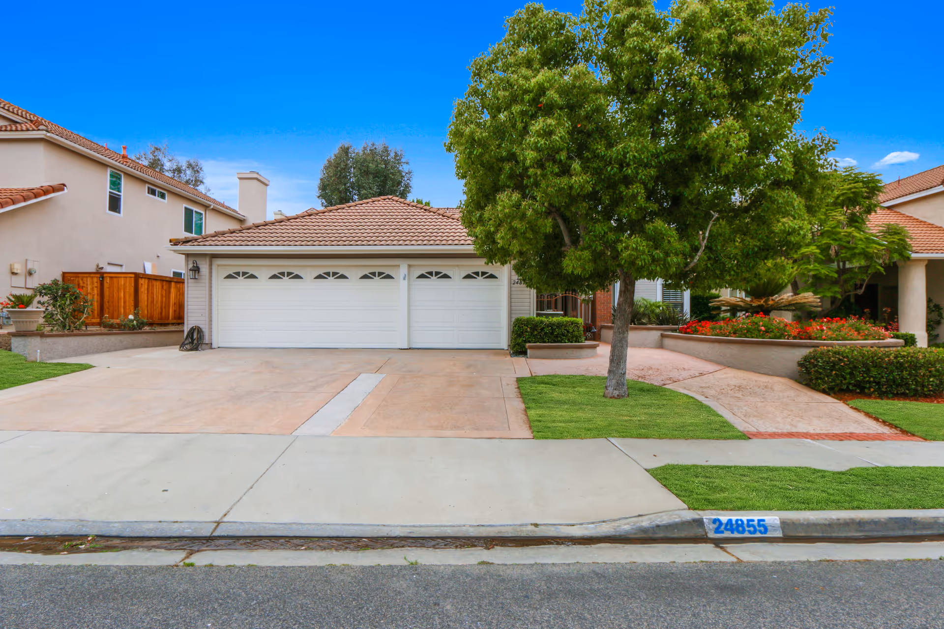 Front exterior view of a single-story house with a three-car garage, a tiled roof, a large tree on the right side, and well-maintained landscaping including green grass and flower beds.