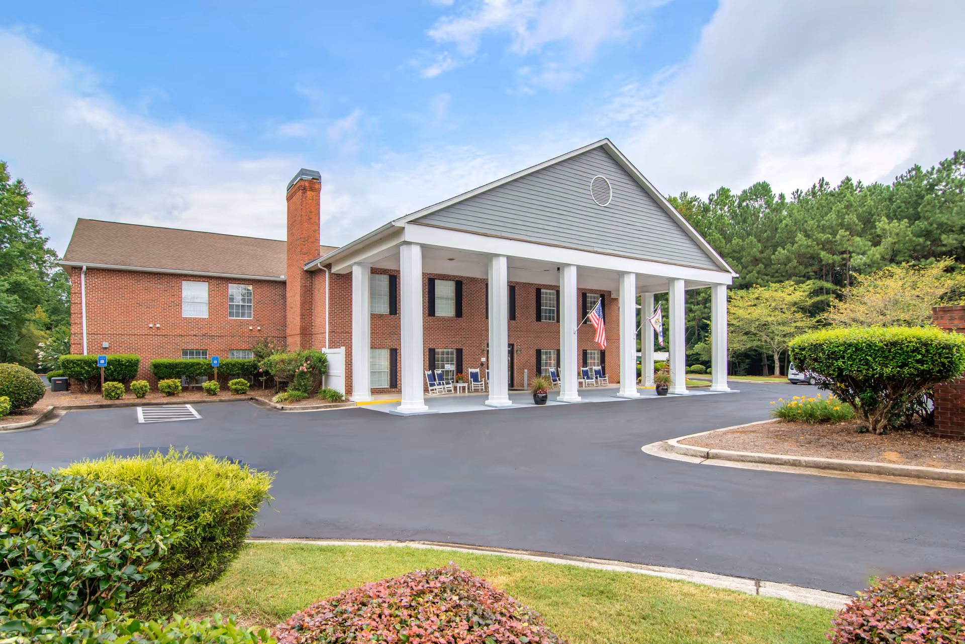 Exterior view of a two-story brick building with white columns supporting a covered entrance. There are American flags hanging from the columns and several rocking chairs on the porch. The building is surrounded by neatly trimmed bushes and trees under a partly cloudy sky.