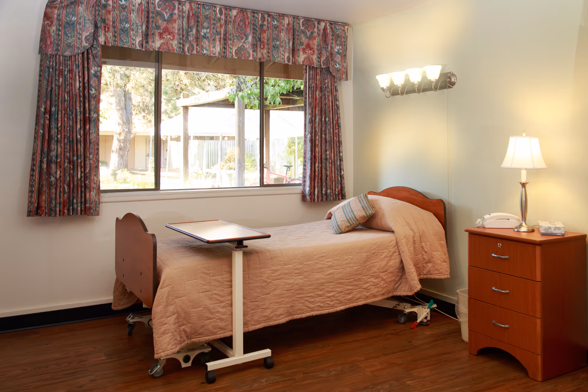 A single bed with a pink quilt, overbed table, bedside dresser and lamp in a well-lit resident bedroom with a large window and patterned curtains.