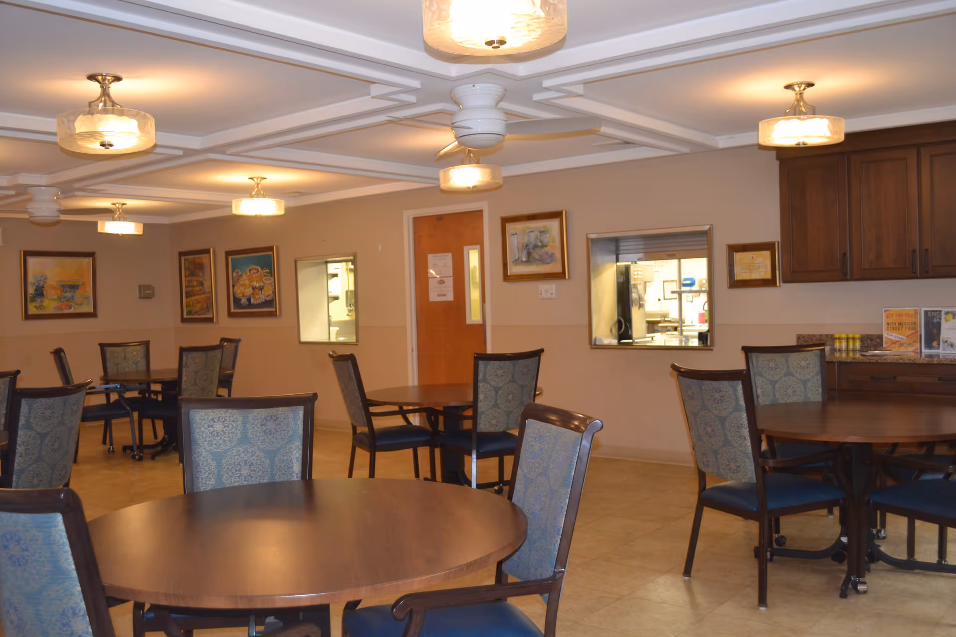 Interior view of a dining room with several round wooden tables and chairs with patterned upholstery. The walls are decorated with framed paintings, and there are windows looking into a kitchen area. The ceiling has multiple light fixtures and a ceiling fan.