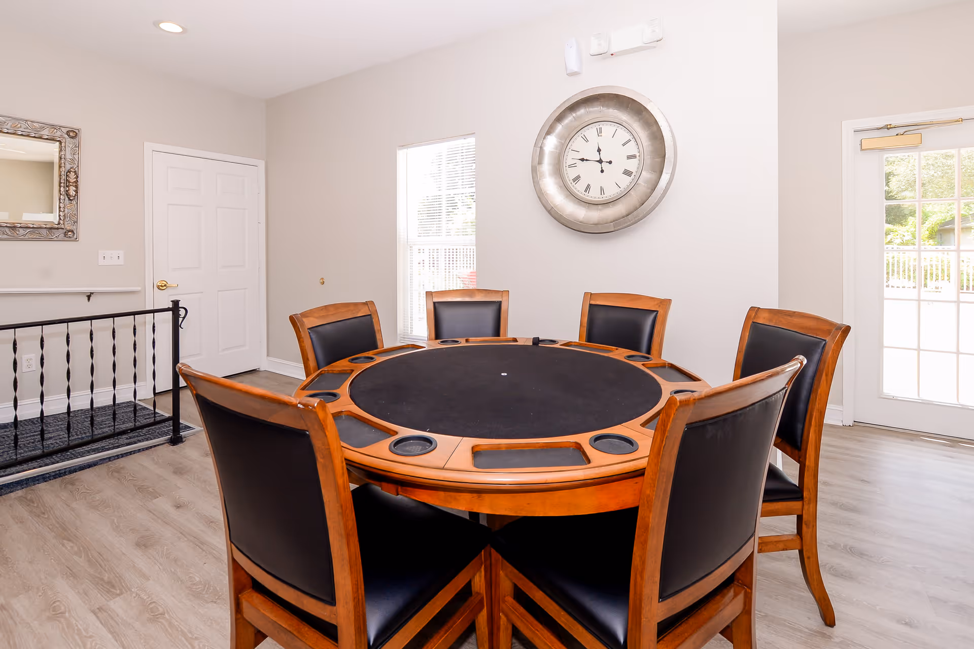 A bright room with a round wooden table surrounded by six wooden chairs with black cushions. The table has built-in cup holders and a black felt playing surface. On the wall behind the table is a large round clock with Roman numerals. There is a window and a glass door letting in natural light, and a white door and a decorative mirror are visible on the left side of the room. The floor is light-colored wood.