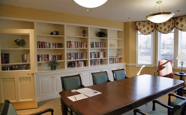 Sunlit meeting room with a long wooden table surrounded by green-upholstered chairs and built-in bookshelves along one wall.